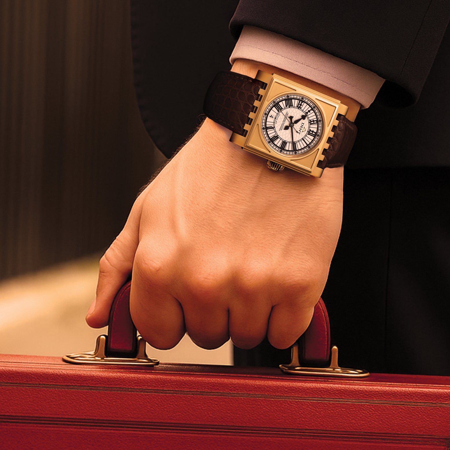 Hand wearing a gold Dent London wristwatch with a leather strap on a red case.