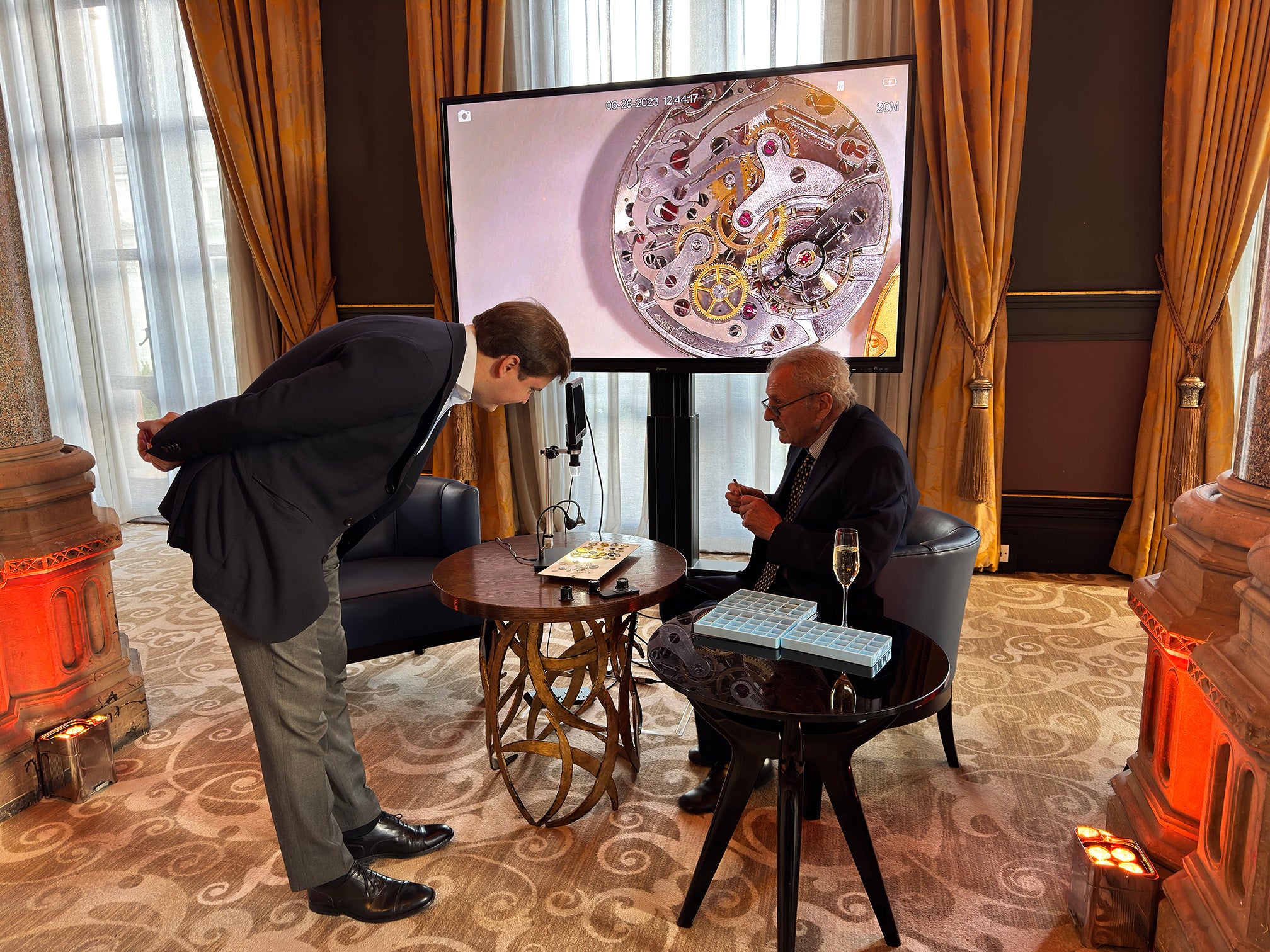 Dent London watchmaker in a formal setting discussing a watch mechanism displayed on a screen.